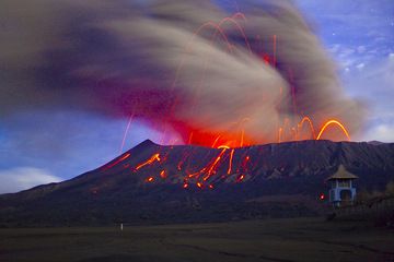 Dense nubi di cenere sono prodotte da esplosioni di tipo vulcaniano. In questo scatto notturno a lunga esposizione, la nuvola di cenere è sfocata sullo sfondo del paesaggio illuminato dalla luna. Il tempio indù di Bromo in primo piano. (Photo: Tom Pfeiffer)