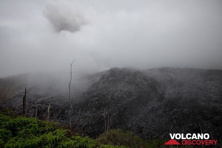 At the rim, fog obscures much of the view, but the dark menacing mass of the lava dome is visible, and an eruption plume can be guessed rising from its center. (Photo: Tom Pfeiffer)