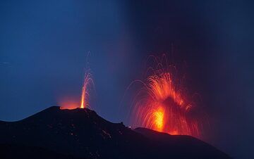 An eruption at the western vent with abundant dark ash occurs while a candle-like stronger spattering or small eruption is seen from the eastern cone. (Photo: Tom Pfeiffer)
