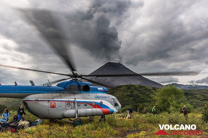 The helicopter keeps the engine running while we unload everything to return as soon as possible, before the weather worsens; fortunately, the rotor blades are at safe height. Karymsky volcano in the background emits a dense steam plume, while the sky is already covered with dense clouds. (Photo: Tom Pfeiffer)