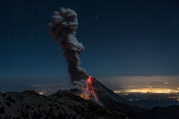 El penacho de ceniza de la erupción se levanta cerca de verticalmente hacia el cielo nocturno con luces de la ciudad de Colima en el fondo. (Photo: Tom Pfeiffer)