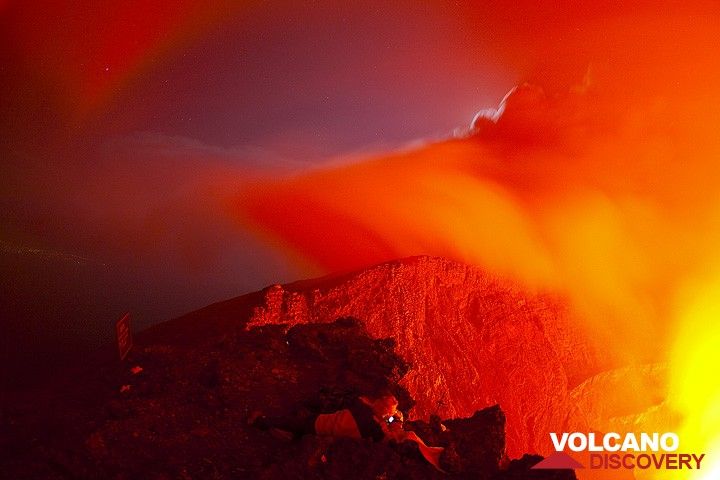 Nyiragongo volcano at night: Gilles in his favorite position, watching the fascinating movements of the lava lake at night for hours. The lights of Goma are visible in the far left. (Photo: Tom Pfeiffer)
