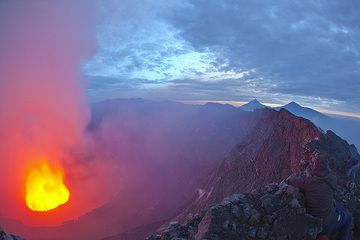 Morning comes fast! A last photo of the lava lake in its giant crater, before fumes become too dense. Jean Michel, at the lower right, merges with the ground. Mikeno and Karisimbi in the background. (Photo: Tom Pfeiffer)