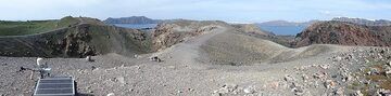 Panoramablick auf das zentrale Gebiet von Nea Kameni mit Eruptions- und Explosionskratern, Überwachungsgeräten (vorne links) und einem rot oxidierten Lavadom und -fluss (rechts). Im Hintergrund sieht man diese Caldera mit (von links nach rechts) der Insel Therasia und den weißen Dörfern Oia und Fira auf der Insel Thera. (Photo: Ingrid Smet)