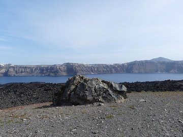 Große Brotkrustenbombe auf Nea Kameni vor einem dunkelgrauen bis schwarzen blockigen Lavastrom. Im Hintergrund sind die Caldera-Klippen im Zentrum von Thera und der Berg Profitis Ilias (rechts) zu sehen. (Photo: Ingrid Smet)