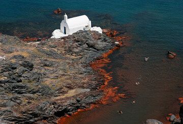 Agios Nikolaos chapel on Palea Kameni island, one of the two historic volcanic islands in the center of the Santorini caldera (Greece). The sea around the island with its small bays is a popular destination for boat cruises - swimmers enjoy the warm waters of the red iron bay.
The orange stain of the rocks on the shore is the result of iron oxide deposit from an active hydrothermal vent system at the shallow sea floor around the Kameni islands. It releases heated, iron-rich waters and deposits a layer of iron-mud on the sea floor. (Photo: Tom Pfeiffer)