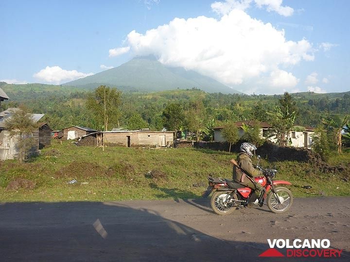Tag 7 – Unser Motorrad-Begleiter aus dem Virunga-Nationalpark begleitet uns auf dem Rückweg nach Goma, mit dem Vulkan Mikeno im Hintergrund (Photo: Ingrid Smet)