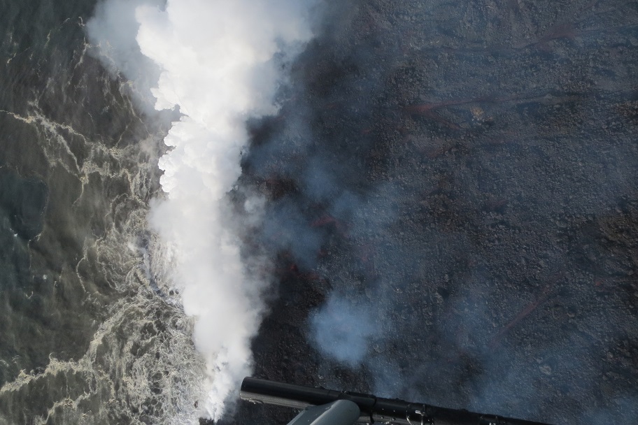 The helicopter hovers above the ocean entry on May 20, 2018, around 6:45 AM HST. Several braided lava channels (red) are visible on the right. The white plume is "laze," which forms when hot lava hits the ocean sending hydrochloric acid and steam with fine glass particles into the air. (HVO/USGS)