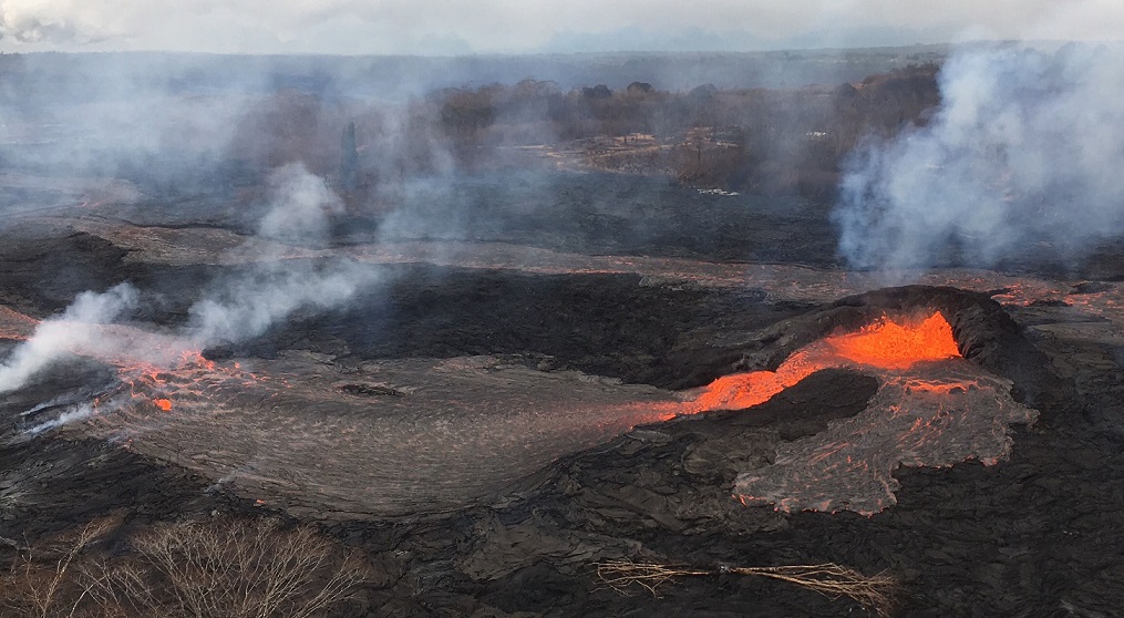 : Activity at fissure 6 on the morning  of 25 May, 2018. Lava fountains have built a small spatter cone (black mound) from which lava was spilling out onto the surface and flowing into a small pond (left of the cone). (HVO/USGS)