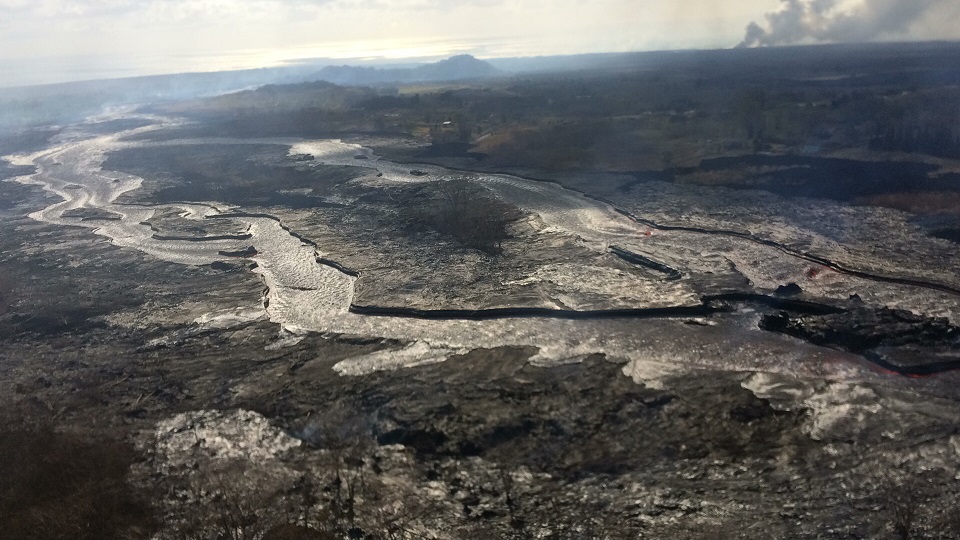 Minor amounts of lava that briefly spill over the lava channel levees are recognisable  as the shiny gray lobes along the channel margins. Image taken during a 17 June, 2018, morning overflight, view to the east, with the plume in the upper right showing the location of the ocean entry. (HVO/USGS)
