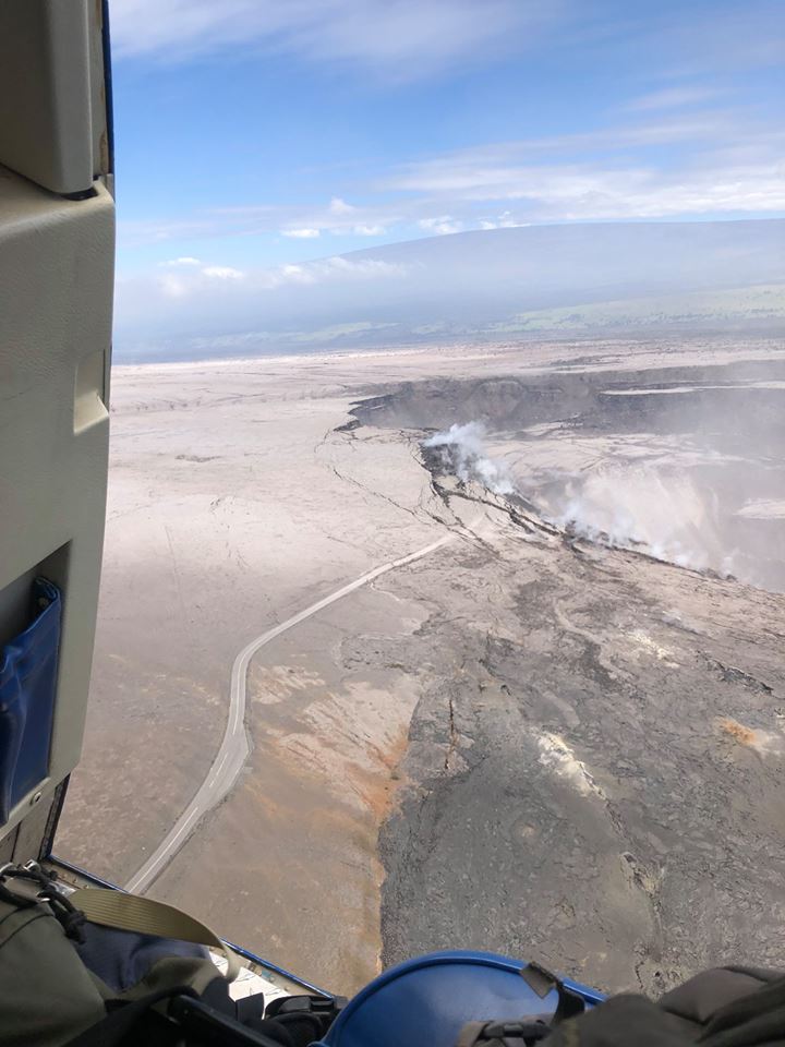 : A helicopter overflight on June 19 confirms that the Overlook parking area (closed since 2008) has largely slumped into the crater of Halema‘uma‘u. The image view is to the northwest and the road beneath the helicopter leads to the former parking area in the center right. (HVO/USGS)